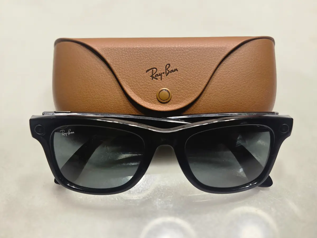 Black Ray-Ban Meta smart glasses with small camera openings in the frame corners, in front of a brown leather charging case