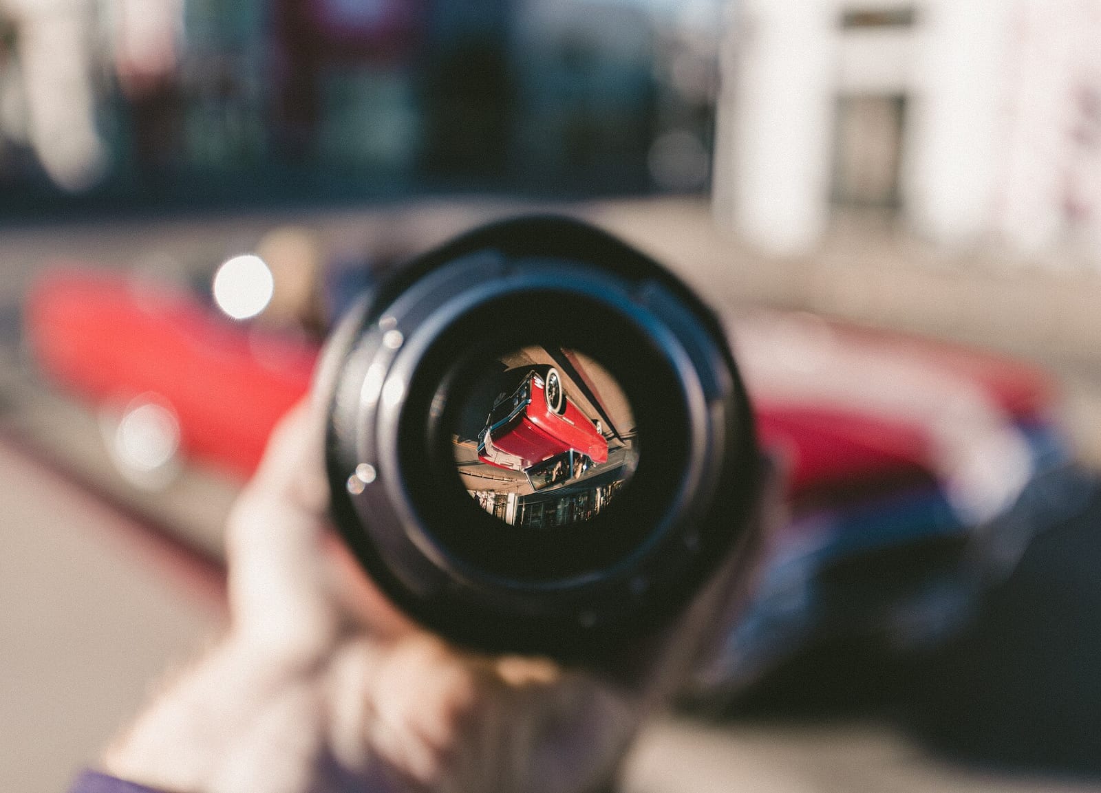 A hand holding a camera lens that reflects a red car on the street