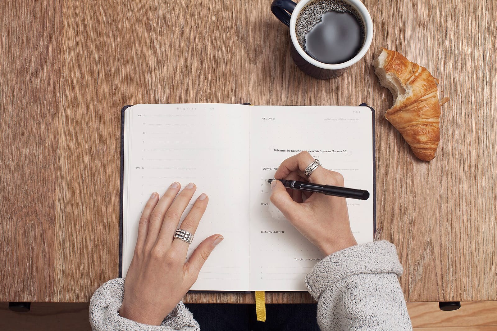 A person writing in a notebook next to a cup of coffee and a croissant