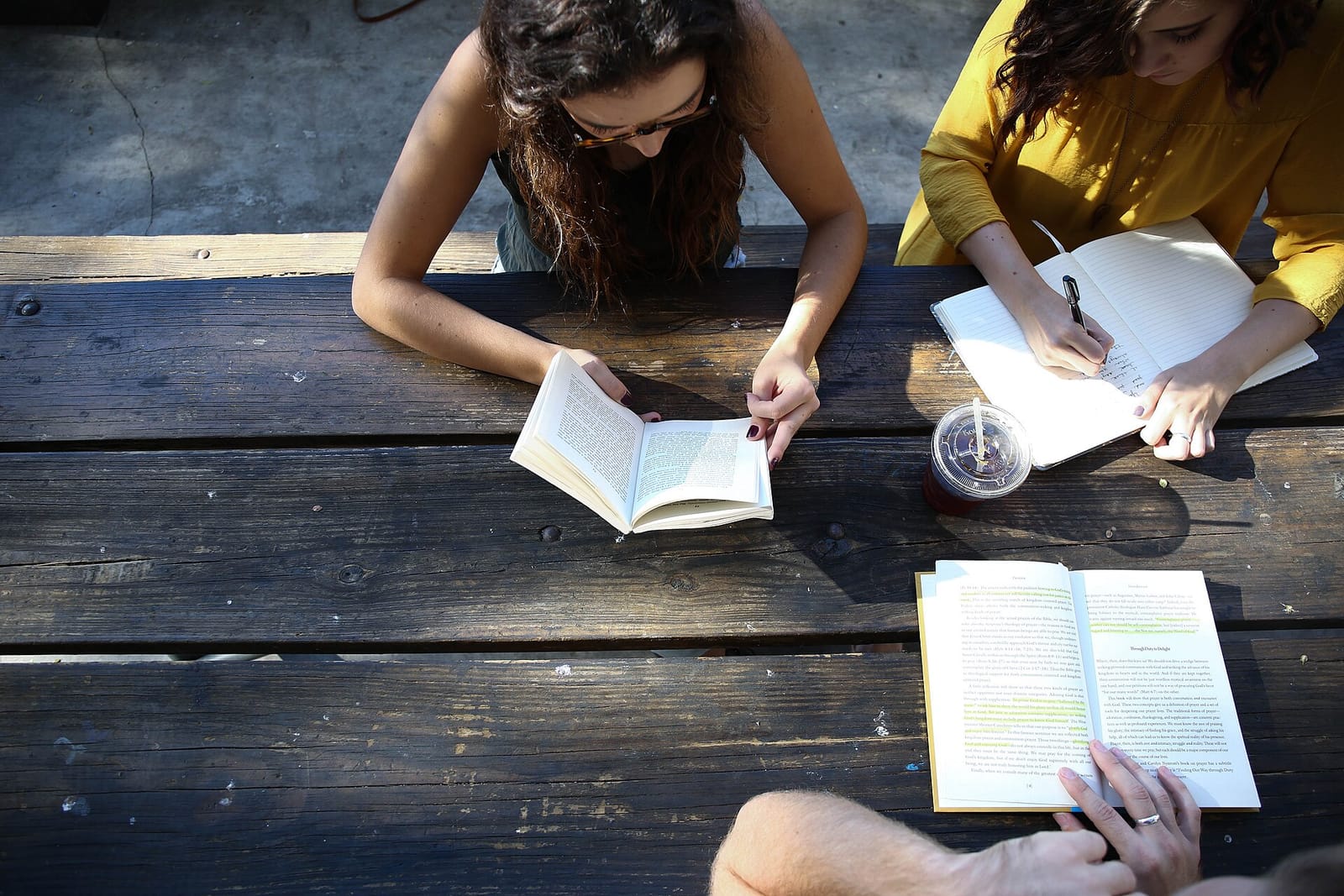 Three people reading and annotating books with highlighters at a wooden table