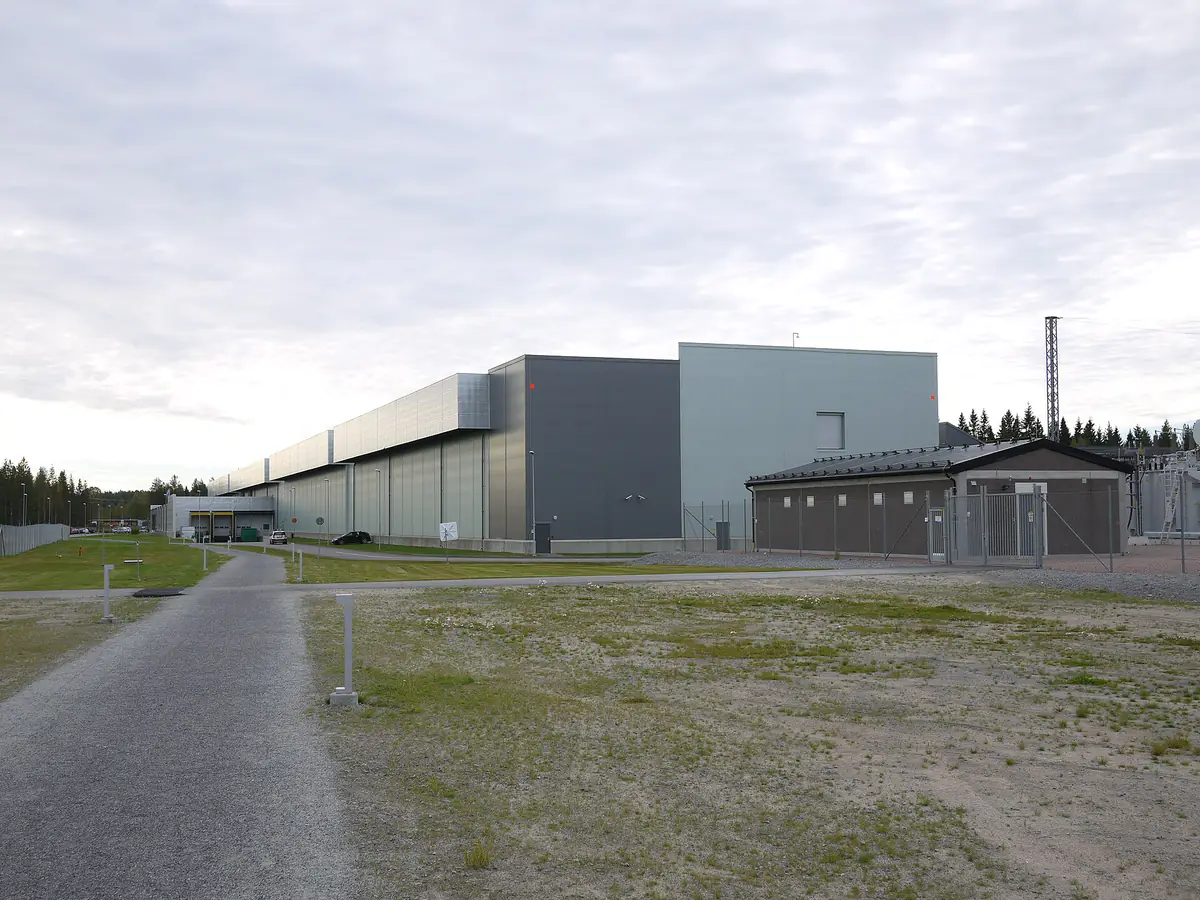 Long, low grey data center halls beside a gravel path and a small fenced building under an overcast sky