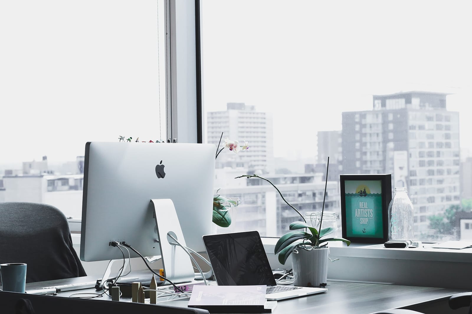 Office desk by a window with an iMac, a laptop and a plant
